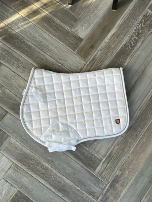 Vitt schabrak Trinity quiltad - Bara använd ett fåtal gånger!💓Säljer ett vitt schabrak från Trinity med quiltat rutmönster och broderad logga. Schabraket har förstärkta partier vid sadelgjorden och praktiska fästen. Passar perfekt till ridning och ger en clean look till din häst. 