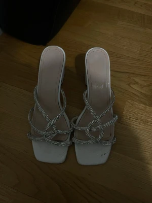 Vita sandaler med strass och klack - Säljer ett par vita sandaler med fyrkantig tå och blockklack. Remmarna är dekorerade med glittrande strass som ger en lyxig känsla. Perfekta för dig som vill sticka ut lite extra. Materialet är syntet och sandalerna har öppen tå.