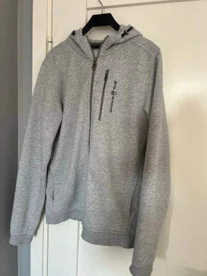 Grå hoodie från Sail racing med dragkedja - Snygg grå hoodie från Sail racing med dragkedja framtill och bröstficka med logga. Tröjan har huva med dragsko och två sidofickor. Tillverkad i mjukt bomullsmaterial, perfekt för chill eller träning. Enkel och stilren design med liten logga även på huvan.