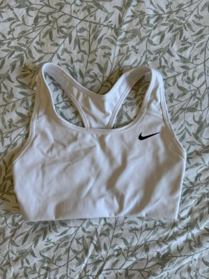 Vit sport-bh från Nike - Snygg vit sport-bh från Nike med klassisk brottarrygg och svart Nike-logga på bröstet. Tillverkad i stretchigt funktionsmaterial som ger bra stöd vid träning. Perfekt för gymmet, löpning eller andra sportaktiviteter 🩷