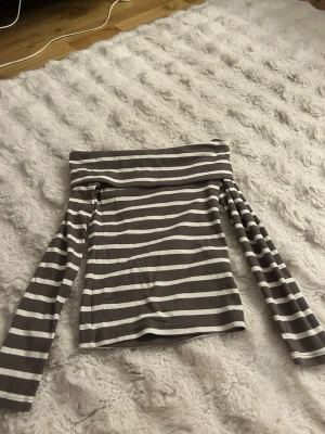 Randig off shoulder topp - Säljer en snygg off shoulder topp med grå och vita horisontella ränder. Toppen har långa ärmar och en bred kant upptill som ger en cool, avslappnad look. Perfekt att styla med jeans eller kjol för en trendig outfit.