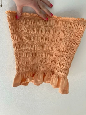 Persikofärgad smockad tubtopp - Supersöt persikofärgad tubtopp med smockad resår och volangkant nedtill. Toppen är axelbandslös och har en stretchig passform som sitter snyggt på kroppen. Perfekt för varma dagar och enkel att styla med jeans eller kjol.