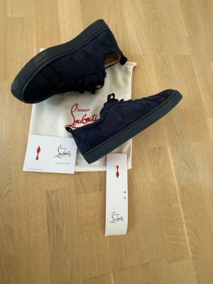Mörkblåa sneakers från Louboutin - Snygga mörkblåa sneakers från Christian Louboutin i mocka med matchande mörkblå sula. Låg modell med ton-i-ton broderad logga på hälen och klassisk siluett. Skorna har rund tå och skön innersula i läder. Perfekta för dig som vill ha en clean och exklusiv look.