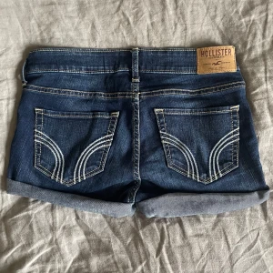 Lågmidjade jeansshorts från Hollister - Snygga mörkblå jeansshorts från Hollister med klassiska bakfickor och uppvikta benslut. Shortsen har normal passform och är perfekta för varma dagar. Tydliga kontrastsömmar och läderpatch med logga bak. Storlek W23. Midjemått rakt över: 36, men stretchiga!!🩷