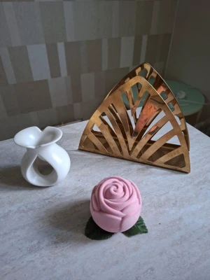 Dekorationspaket med ros och hållare - Säljer ett dekorationspaket med en ljusrosa ros i tyg, en vit keramikljushållare med modern form och en triangulär servetthållare i guldmetall med art deco-mönster. Perfekt för att piffa upp hemmet med unika detaljer.