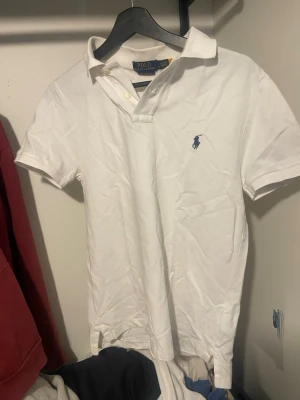 Vit pikétröja från Polo Ralph Lauren - Klassisk vit pikétröja från Polo Ralph Lauren med blå logga broderad på bröstet. Tröjan har korta ärmar, krage och knappar vid halsen. Modellen är custom slim fit och tillverkad i bomull för en bekväm känsla. Storlek S. Skriv vid frågor!