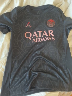 PSG x Jordan svart matchtröja M - Snygg svart PSG matchtröja från Jordan med diskret mönster, rosa loggor och Qatar Airways-tryck på bröstet. Tillverkad i Dri-FIT-material som andas, perfekt för träning eller match. Rund hals och korta ärmar för sportig look. 