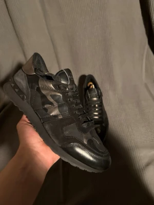  Valentino rockrunner  - Snygga svarta sneakers från Valentino med camo-mönster i olika nyanser av svart och grått. Skorna har en låg silhuett, svarta skosnören och detaljer i både läder och textil. Perfekta för dig som vill ha en stilren men ändå unik look.