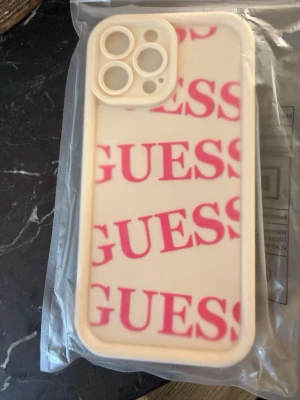 Guess mobilskal i beige och rosa - Snyggt mobilskal från Guess till iPhone 13 Pro Max. Skalets bas är beige med rosa GUESS-logga tryckt flera gånger över hela baksidan. Tillverkat i tåligt plastmaterial och har förstärkt ram runt kameran för extra skydd. Perfekt för dig som vill ha en trendig look på mobilen.