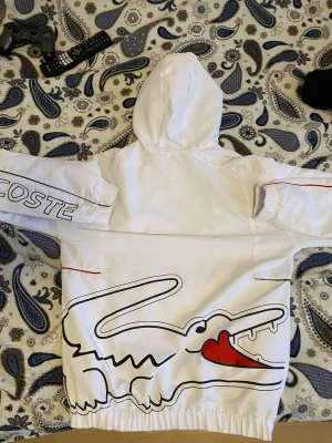 Lacoste zip hoodie - Säljer en vit zip hoodie från Lacoste med stor krokodil-illustration och röd detalj på ryggen. Ärmarna har elastiska muddar och trycket 'LACOSTE' på ena ärmen. Hoodien har huva och cool streetwear-vibe.