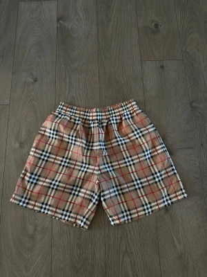 Beige rutiga shorts från Burberry - Snygga beige shorts från Burberry med klassiskt rutigt mönster i svart, vitt och rött. Shortsen har elastisk midja för skön passform och är perfekta för sommaren. Tillverkade i ett lätt och bekvämt material.