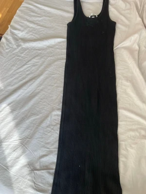 Svart ribbad långklänning - Säljer en svart ribbad långklänning med breda axelband och rund halsringning. Klänningen har en tight passform och är tillverkad i ett stretchigt material som sitter snyggt på kroppen. Perfekt för dig som gillar en enkel men stilren look.