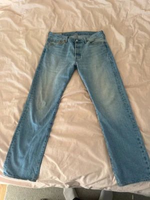 Levi's 501 ljusblå raka jeans - Klassiska Levi's 501 jeans i ljusblå denim med rak passform och fem fickor. Jeansen har knappgylf och den ikoniska läderpatchen bak i midjan. Perfekta för en avslappnad och tidlös stil.