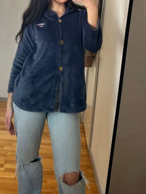 Mörkgrå fleecekofta med huva - Mysig mörkgrå kofta från Christine Bernadotte i mjuk fleece. Har huva, stora träknappar och broderad text 'Fabulous Girl' på bröstet. Perfekt för kyliga dagar och enkel att soppa över en t-shirt. Långärmad och med fickor på sidorna.