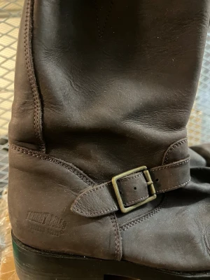 Bruna läder cowboy boots från Johnny Bulls - Snygga bruna cowboy boots från Johnny Bulls i äkta läder. Bootsens design har ett robust spänne i metall på sidan och en slitstark sula som är oljeresistent. Perfekta för dig som gillar westernstil och vill ha något som sticker ut.