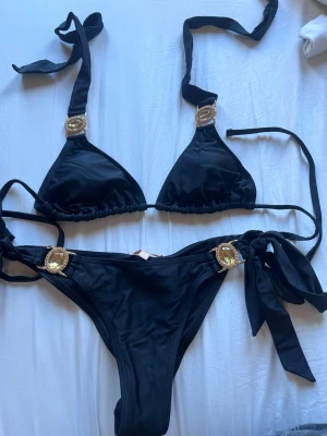 Svart bikini från Moda Minx med gulddetaljer - Snygg svart bikini från Moda Minx med guldfärgade dekorativa detaljer på både överdel och underdel. Bikinin har knyt i sidorna och runt nacken för justerbar passform. Perfekt för stranden eller poolen! Aldrig använd 
