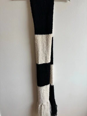 Randig svartvit halsduk med fransar - Säljer en lång, mjuk halsduk i svart och vitt med breda blockränder. Halsduken har fransar i båda ändar och är stickad i ett fluffigt material🧣