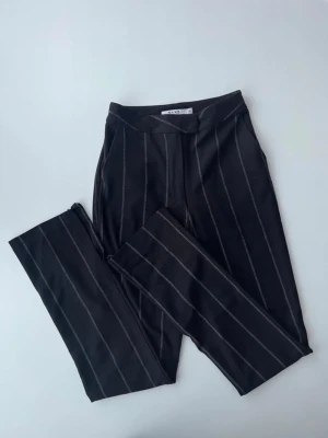 Svarta kostymbyxor med ränder från Nakd  - Snygga svarta kostymbyxor från Nakd med tunna vita kritstrecksränder. Byxorna har rak passform och klassisk midja. Perfekta för en stilren och trendig look. Materialet känns mjukt och följsamt.