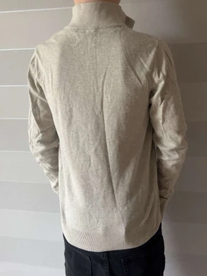Beige halvzip tröja med hög krage - Snygg beige tröja med halv dragkedja och hög krage. Tröjan har långa ärmar och ribbade muddar vid ärmslut och nederkant. Perfekt för en avslappnad men stilren look. Materialet känns mjukt och bekvämt mot huden. OBS! Barnstorlek 152