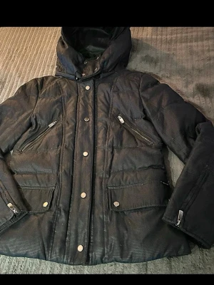 Zara svart puffer jacka med avtagbar huva  - Säljer en svart pufferjacka från Zara med huva och snygga detaljer som dragkedjor och tryckknappar. Perfekt till höst/vinter – både varm och stilren.  Jackan har flera fickor och en mjuk, lite “läderliknande” struktur som ger ett exklusivt look.  Skick: använd men fortfarande i bra skick. Lite normalt slitage finns (se bilder), inget som påverkar användning.  Passar till allt – streetwear, basic outfits eller uppklätt. Längd - ca 63 cm Axel till axel - ca 49 cm Ärmlängd - ca 61 cm