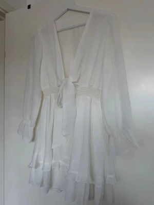 Vit omlottklänning med volanger - Säljer en superfin vit kortklänning med omlott och knyt framtill. Klänningen har långa ärmar med volangdetaljer vid ärmslut och nederkant, samt smock i midjan för en snygg passform. Perfekt för sommarens alla tillfällen! Eller till strudent och skolavslutning 