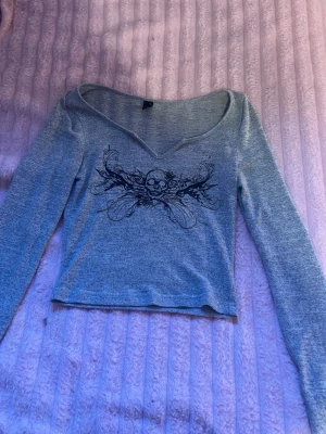 Grå långärmad topp med dödskalle - Grå croppad långärmad topp med v-ringning och svart dödskalletryck framtill. Tunn och mjuk i materialet, perfekt för en edgy stil. Snygg att matcha med jeans eller kjol.