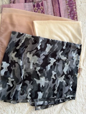 Sjalar i olika färger och mönster - Fyra sjalar i olika stilar: en med svart, grå och vit camouflage, en ljusbeige, en ljusrosa och en lila med etniskt mönster. Perfekta för att addera färg och personlighet till din outfit. Materialet känns mjukt och lätt, troligen polyester eller viskos.
