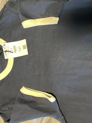 Svart t-shirt med gula detaljer Bershka - Snygg svart t-shirt från Bershka med kontrasterande ljusgula muddar vid halsringning och ärmslut. Klassisk passform och rund hals. Perfekt för en avslappnad och trendig stil. Tillverkad i mjuk bomull för skön känsla hela dagen.