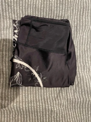 Svarta leggings från Anarchy Apparel - Snygga svarta leggings från Anarchy Apparel med vit blommig print och stor logga längs benet. Tillverkade i stretchigt och mjukt material som sitter skönt på kroppen. Perfekta för dig som vill ha något unikt och sportigt i garderoben.