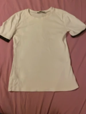 Vit t-shirt med glittriga ärmslut - Säljer en vit t-shirt med korta ärmar och rund hals. Ärmsluten har en snygg svart kant med glittriga detaljer som ger en extra touch. Perfekt för dig som vill ha något enkelt men ändå med lite edge.