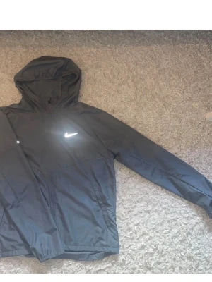 Nike svart träningsset med huva - Säljer ett svart träningsset från Nike med jacka och byxor. Jackan har huva, dragkedja och reflekterande Nike-logga på bröstet. Byxorna har resår i midjan och reflekterande logga på benet. Perfekt för löpning eller gym. Materialet är lätt och andas.