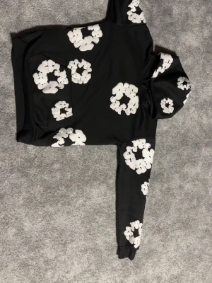 Svart hoodie med vita blommor - Säljer en svart hoodie med stora vita blommönster över hela plagget. Hoodien har en klassisk huva och ribbade muddar vid ärmsluten. Materialet känns mjukt och bekvämt, perfekt för en avslappnad stil. Inga synliga loggor eller märken.