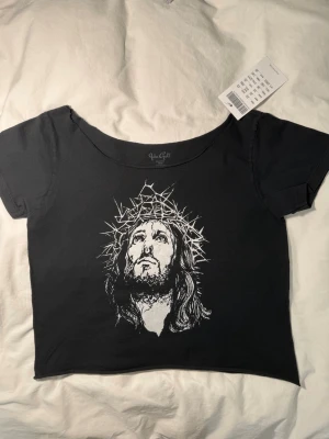 Svart croppad t-shirt John Galt / Brandy Melville - Svart croppad t-shirt från John Galt med Jesus. T-shirten har korta ärmar och en bred, rund halsringning. Tillverkad i 100% bomull för en mjuk och bekväm känsla. Perfekt för dig som gillar edgy och unik stil.