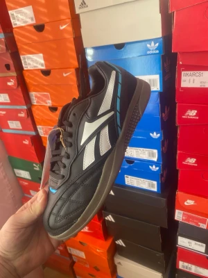 Svarta Reebok sneakers med vit logo - Snygga svarta Reebok sneakers med vit logga på sidan och blå detaljer. Skorna har en låg profil, klassisk gummisula och ovandel i läder och syntet. Perfekta för dig som gillar retrostil och sportig look.