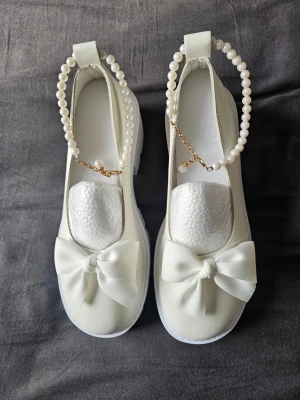 Vita ballerinaskor med pärlband - Säljer ett par cremefärgade/vita ballerinaskor med chunky sula och stora rosetter framtill. Skorna har dekorativa pärlband med guldfärgade detaljer runt ankeln som ger en söt och unik look. Trendig design.högkvalitativt konstläder, och halkfri gummisula. Köpta för 398 kr. på Amazon.  Helt nya, aldrig används, Det står stl 43, men i verkligheten är det stl.41.  Priset kan diskuteras 
