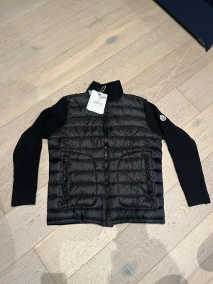 Svart cardigan från Moncler - Snygg svart cardiganfrån Moncler med stickade ärmar och hög krage. Västen har två dragkedjeförsedda fickor framtill och Moncler-logga på ena ärmen. Framsidan är quiltad och fylld med dun för extra värme, medan ärmarna är ribbstickade för en cool kontrast.