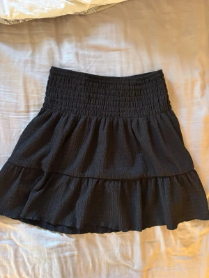 Svart volangkjol🫶🏻 - Säljer en svart kortkjol med bred resår i midjan och volangdetaljer. Kjolen har en luftig och flowy känsla, perfekt för varmare dagar. Materialet känns mjukt och bekvämt mot huden. Enkel att matcha med olika toppar för en snygg look. Så gullig!!🤍🤍