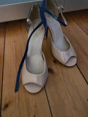 Beige och blåa peep toe pumps - Snygga pumps med öppen tå i beige lack och blåa detaljer. Skorna har en smal klack och justerbart ankelband i blått. Perfekta för dig som vill sticka ut med en cool färgkombination. Märke W syns på innersulan. 