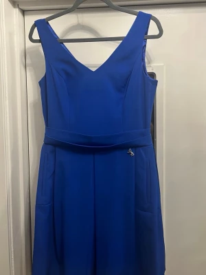 Kornblå ärmlös klänning med v-ringning - Snygg kornblå klänning med v-ringning både fram och bak, markerad midja och dekorativ dragkedja i ryggen. Klänningen är ärmlös och har fickor i sidorna. Perfekt för dig som vill ha en stilren och modern look. 