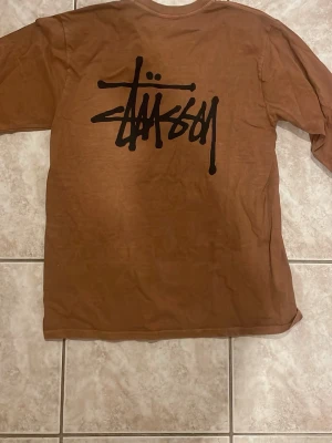 Brun långärmad Stüssy tröja - Säljer en brun långärmad tröja från Stüssy med ikonisk svart logga tryckt stort på ryggen och mindre på bröstet. Tröjan har rund halsringning och är gjord i mjukt bomullsmaterial. Perfekt för dig som gillar streetwear och vill ha något stilrent men ändå utstickande.