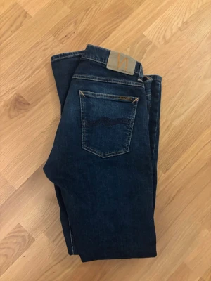 Mörkblå jeans från Nudie Jeans - Säljer ett par klassiska mörkblå jeans från Nudie Jeans med fem fickor och snygga kontrastsömmar. Jeansen har en normal passform och är tillverkade i mjuk bomull. Perfekta till alla dina outfits!