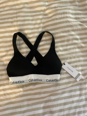 BH Calvin Klein  - Snygg svart sport-bh från Calvin Klein med bred resår i vitt och logga runt om. Bh:n har korsade axelband i ryggen och är tillverkad i mjukt stretchigt material för extra komfort.