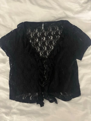 Svart spetsblus från Pieces - Snygg svart blus från Pieces i genombruten spets med volangkant längs den djupa v-ringningen. Kortärmad modell med knytband framtill. Perfekt för att addera en edgy touch till din outfit. Stl L men passar även mindre