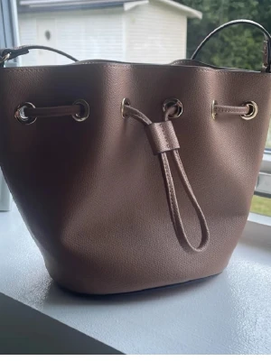 Brun bucket bag med axelrem - Snygg brun bucket bag i konstläder med justerbar axelrem och dragskostängning. Väskan har guldfärgade metalldetaljer och en stilren, modern look. Perfekt för dig som gillar trendiga accessoarer och vill ha plats för det viktigaste.