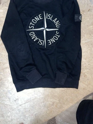 Svart hoodie från Stone Island - Säljer en svart hoodie från Stone Island med stort vitt kompass-tryck och texten 'Stone Island' på bröstet. Klassisk huva och avslappnad passform, perfekt för dig som gillar streetwear och vill ha en stilren look.Storlek S/M