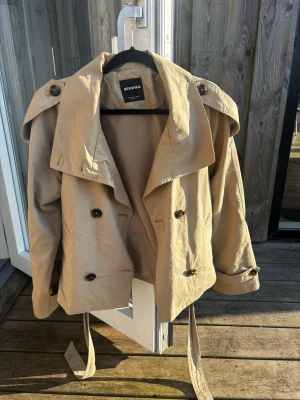 Kort beige trenchcoat med knytskärp - Stilren kort trenchcoat i beige med dubbelknäppning, höga axelklaffar och knytskärp i midjan. Jackan har stora mörka knappar, hög krage och justerbara ärmslut. Perfekt för dig som vill ha en trendig och klassisk look. Materialet ser ut att vara bomull eller bomullsblandning. Den här gröna nyanser men är den beiga modellen. 