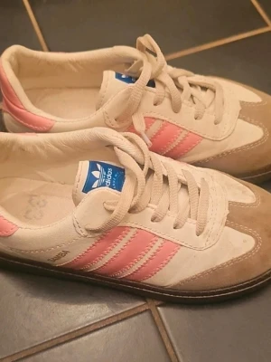 Adidas Samba sneakers rosa/vit - Säljer ett par Adidas Samba sneakers med klassiska rosa ränder, vit ovandel och beige mockadetaljer. Skorna har gummisula, snörning och blå Adidas-logga på plösen. Perfekta för dig som gillar retrostil och vill sticka ut med färg.