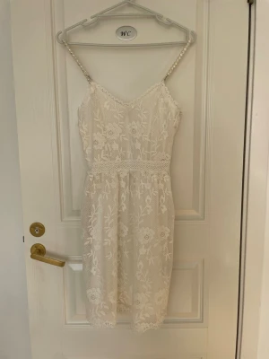 Vit spetsklänning med smala axelband - Söt vit klänning med smala axelband och vackra spetsdetaljer. Klänningen har blommigt mönster, markerad midja och är knälång. Perfekt för sommarens alla tillfällen. Axelbanden är dekorerade med pärlor för extra detalj.