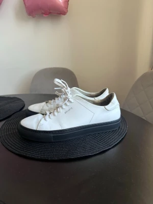 Vita sneakers från Axel Arigato - Stilrena vita sneakers från Axel Arigato med svart sula och klassisk låg design. Skorna är tillverkade i läder och har vita skosnören samt diskret logga på sidan. Perfekta för dig som gillar minimalistisk och clean stil.