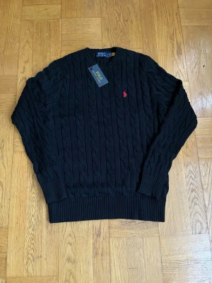 Svart kabelstickad tröja Polo Ralph Lauren - Kabelstickad svart tröja från Polo Ralph Lauren med rund halsringning och ribbade muddar. Klassisk röd logga broderad på bröstet. Tröjan har en tidlös design och är tillverkad i mjukt material som passar perfekt till alla tillfällen.